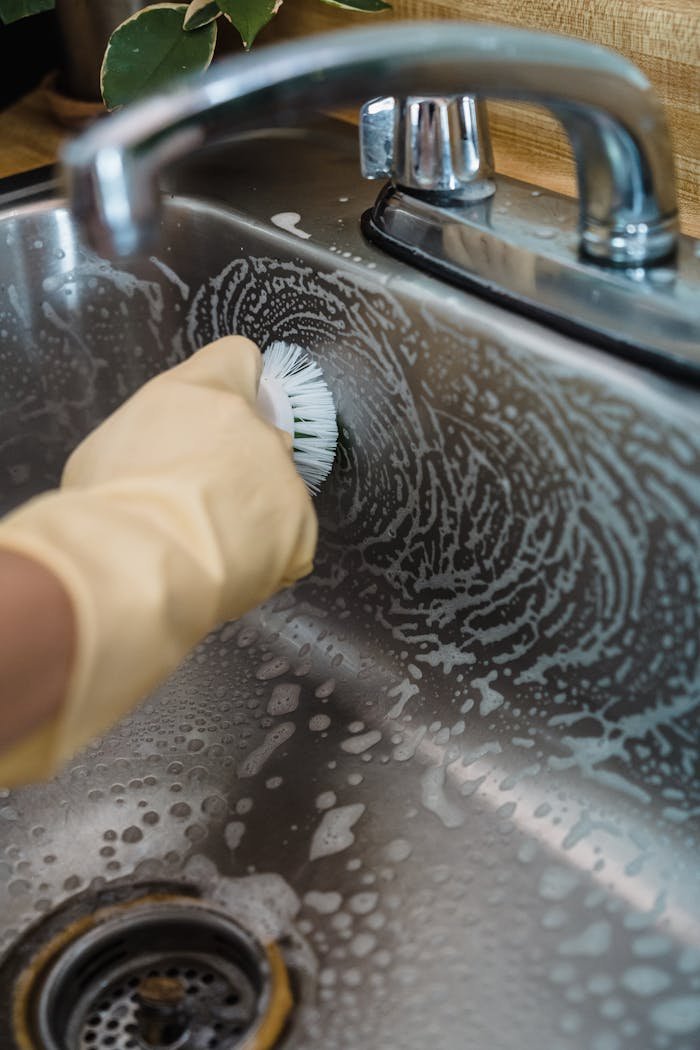 Close-up of hand with glove scrubbing a stainless steel sink with soap and brush.